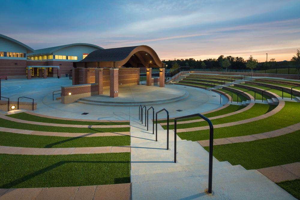 Outdoor amphitheater with tiered seating featuring lawn turf grass, integrated walkways, and a covered stage area in a landscaped public space.